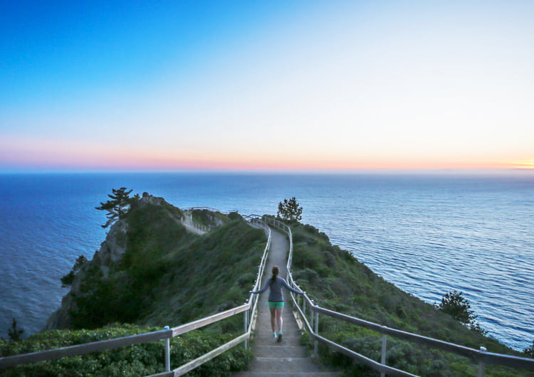 Young woman running along a path out on a rock outcrop along the Pacific Ocean at sunset.